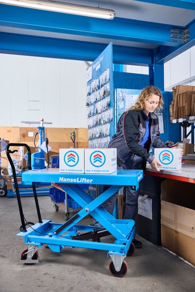 Blaue HanseLifter Scherenhubwagen in einem hellen, organisierten Lager beim Transport von Kartons durch eine Arbeitskraft in Arbeitskleidung.