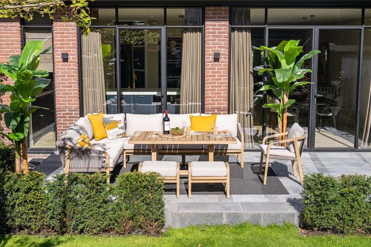 Modernes Gartenmöbel-Set auf einer Steinterrasse mit einem Ecksofa aus Holz, einem Esstisch, zwei Hockern und einem Sessel.