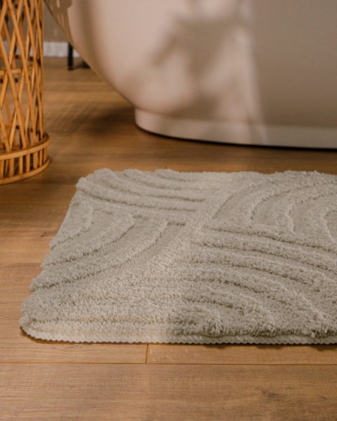 Strukturierte Badematte mit Wellenmuster auf Holzboden neben einer Badewanne und einem Weidenkorb in einem hellen Badezimmer.