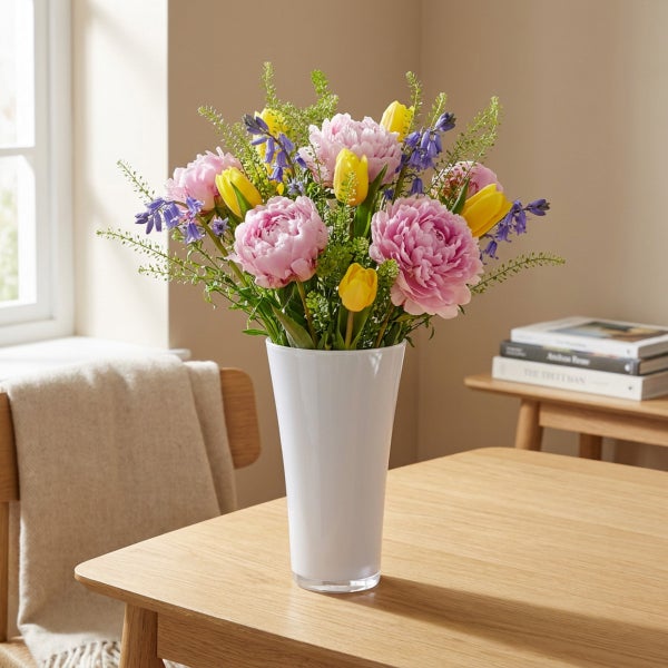Ein Blumenstrauß mit Pfingstrosen und Tulpen in einer hohen weißen Vase auf einem Holztisch in einem hellen Raum.