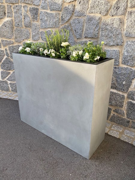Hoher rechteckiger Pflanzkübel in grauer Beton-Optik, bepflanzt mit grünen Pflanzen und weißen Blumen vor einer Steinwand.