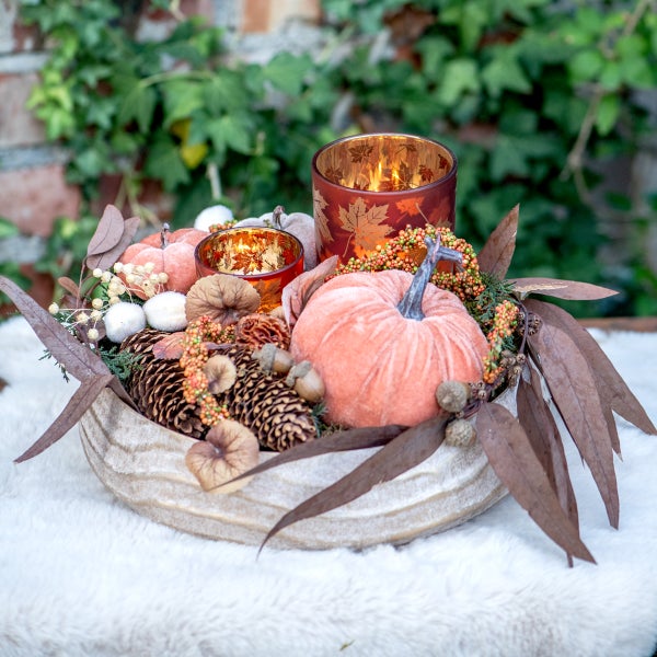 Herbstdekoration in einer strukturierten Schale mit Samtkürbissen, Tannenzapfen, Trockenlaub und zwei brennenden Teelichthaltern mit Blattmotiv.