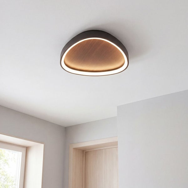 Moderne dreieckige LED Deckenleuchte mit Holzoptik in der Mitte und umlaufendem Lichtring an einer weißen Decke in einem hellen Raum.