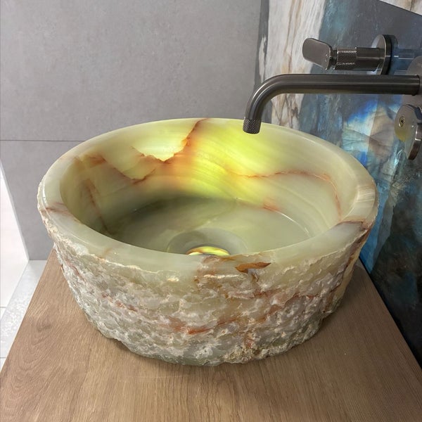 Rundes Aufsatzwaschbecken aus Onyx-Naturstein mit rauer Außenseite und glatter Innenseite, montiert auf einer Holzplatte mit Wandarmatur.