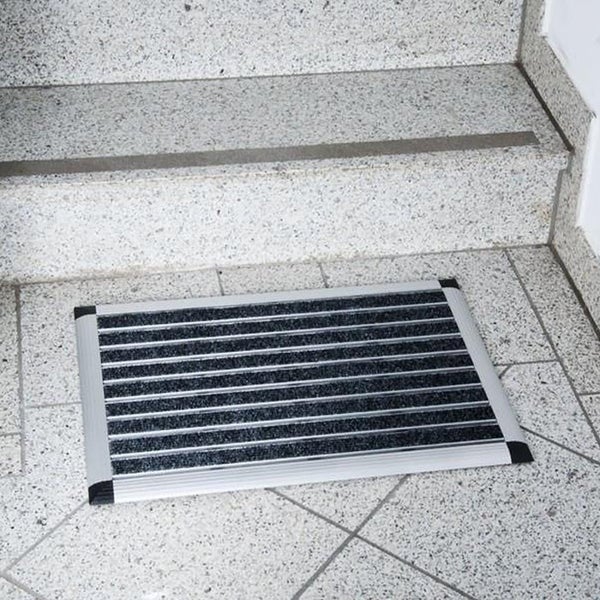 Fußmatte mit Aluminiumrahmen und dunklen Ripseinlagen auf einem Fliesenboden vor einer Treppe.