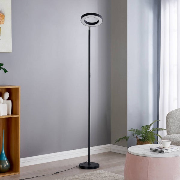 Moderne schwarze LED-Stehleuchte mit ringförmigem Leuchtkopf in einem hellen Wohnzimmer mit grauer Wand und Holzregal.
