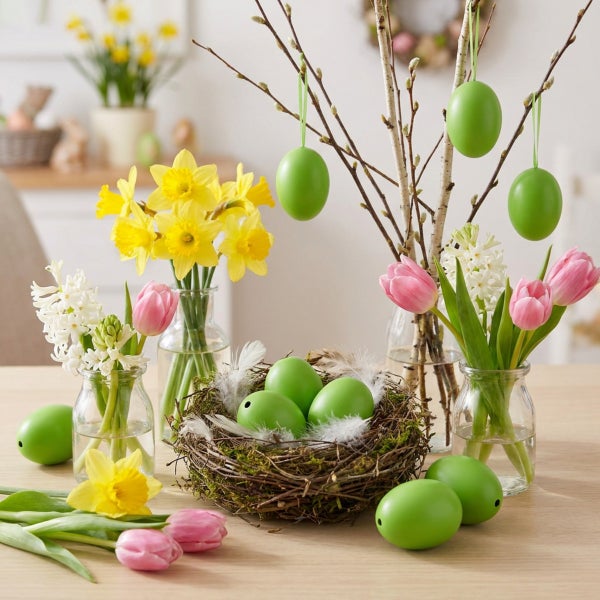 Osterdekoration mit Ostereiern in einem Nest, Zweigen in Glasvasen, Narzissen und Tulpen auf einem Holztisch für eine frühlingshafte Tischdeko.