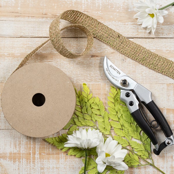 Gartenschere aus Karbonstahl neben einer Rolle Juteband, weißen Blumen und grünen Blättern auf einer Holzoberfläche.