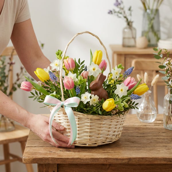 Osterkorb aus geflochtenem Holz mit Frühlingsblumen wie Tulpen, Narzissen und Traubenhyazinthen sowie einer Schokoladenosterhasenfigur.