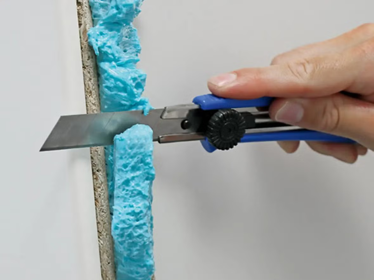 Hand schneidet mit einem Cuttermesser überschüssigen Montageschaum an einer Wandkante ab