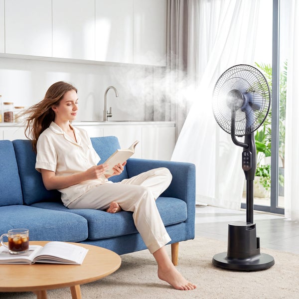 Heller Wohnraum mit einer Frau auf einem blauen Sofa und einem schwarzen Sprühnebelventilator, der feinen Wassernebel zur Kühlung verteilt.