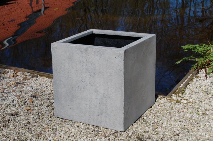 Quadratischer Pflanzkübel in Betonoptik auf Kies am Wasser in einem hellen Garten.
