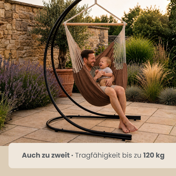 Ein Vater und sein Kind sitzen gemeinsam in einem Hängesessel mit schwarzem Metallgestell auf einer Terrasse in einem Garten.