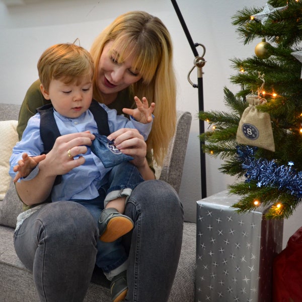 Mutter und Kind sitzen auf einem Sofa neben einem geschmückten Weihnachtsbaum und öffnen gemeinsam ein Säckchen eines Adventskalenders.