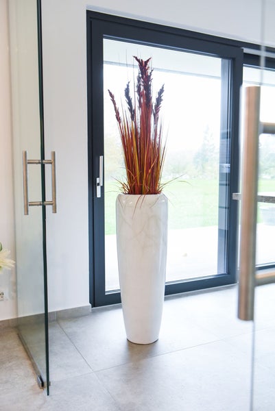 Hohe dekorative Bodenvase in Marmoroptik mit rotbraunen Ziergräsern in einem hellen modernen Raum mit großen Glasfronten.