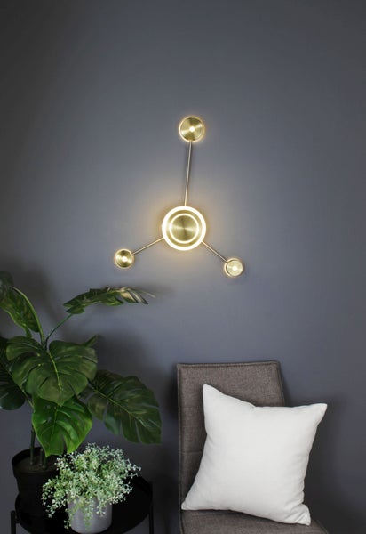 Moderne Wandleuchte aus Metall in Messingoptik mit zentralem Lichtring und drei Armen an einer dunklen Wand neben einer Zimmerpflanze.