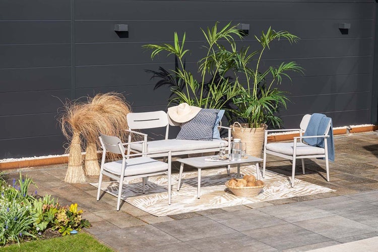 Moderne Garten-Lounge-Gruppe mit Bank, zwei Sesseln und Tisch auf gemustertem Außenteppich, dekoriert mit Pflanzen vor einer dunklen Wand.