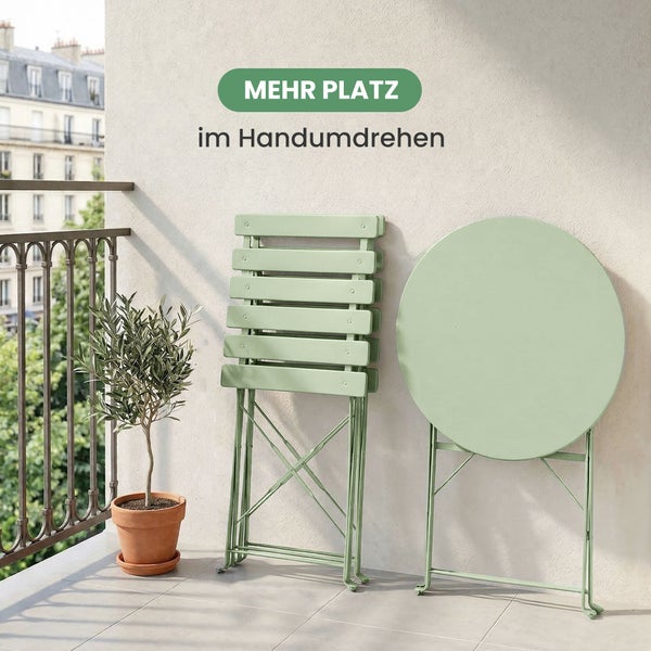 Platzsparende zusammengeklappte Balkonmöbel aus Metall mit rundem Tisch und zwei Klappstühlen an einer Wand auf einem hellen Balkon.
