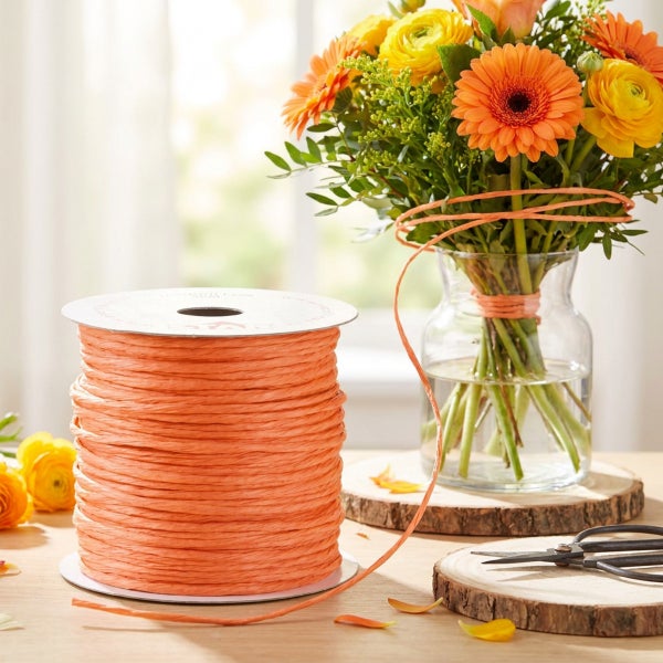 Eine Spule mit orangefarbenem Bastband zum Dekorieren von Blumensträußen, neben einer Glasvase mit frischen Blumen auf Holzscheiben.