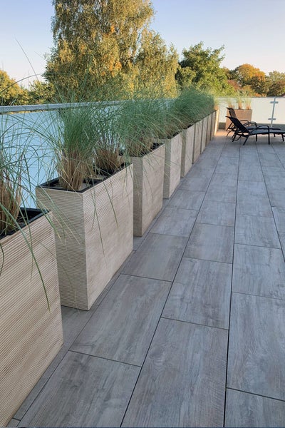 Moderne Dachterrasse mit Bodenfliesen in Holzoptik und einer Reihe hoher, strukturierter beiger Pflanzgefäße mit Ziergräsern.