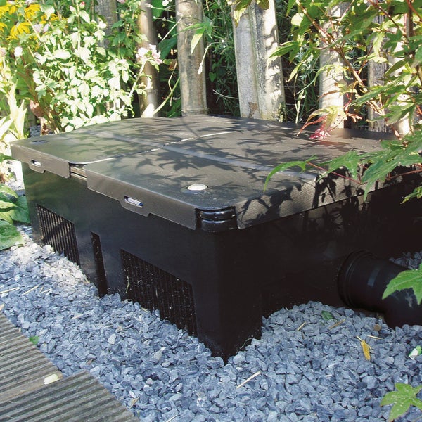 Schwarze Kunststoff-Pumpenkammer im Garten auf grauem Kies platziert, umgeben von Pflanzen und einem Anschlussrohr.