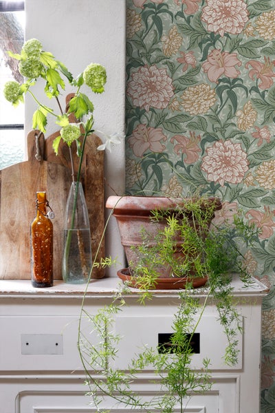 Dekoratives Stillleben mit Blumenmuster-Tapete, Topfpflanze und Vintage-Elementen auf einem weißen Schrank.