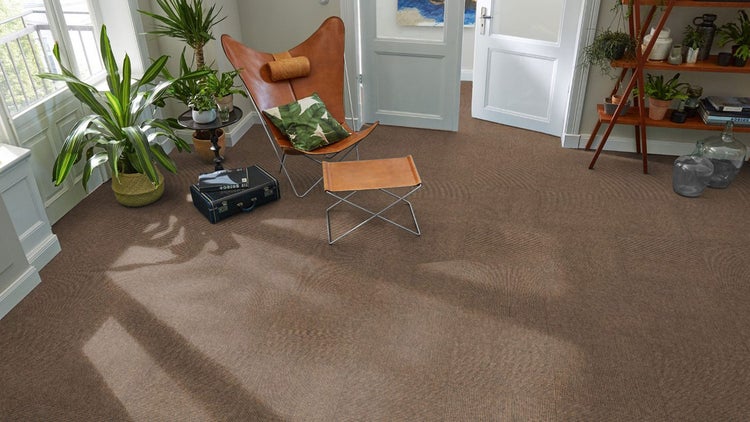 Braune Teppichfliesen mit Struktur in einem hellen Wohnzimmer mit Ledersessel, Hocker und Zimmerpflanzen.