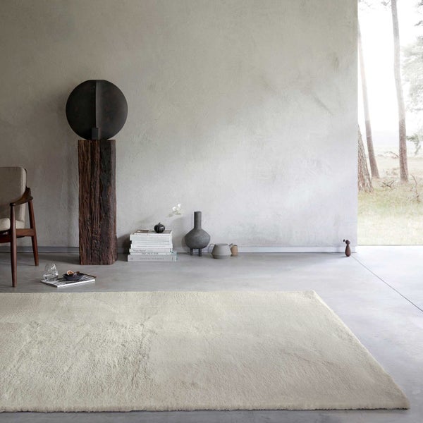 Hellbeiger, weicher Hochflor-Teppich in einem hellen, minimalistischen Wohnraum mit Betonboden, Strukturwand und dekorativen Holz-Skulpturen.