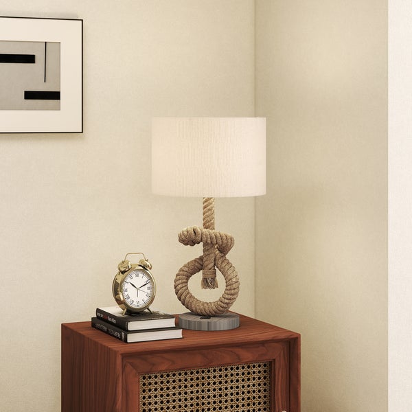 Tischleuchte mit geknotetem Seilfuß und hellem Stoffschirm auf einem Holzschrank neben einem Wecker und Büchern.