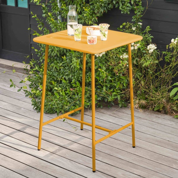 Quadratischer Stehtisch aus gelbem Metall auf einer Holzterrasse mit Gläsern und einer Karaffe vor grünen Pflanzen.