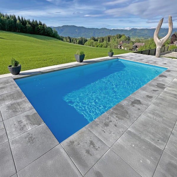 Rechteckiger Außenpool mit blauem Wasser und grauen Terrassenplatten aus Stein vor einer Kulisse aus grünen Wiesen und Bergen.
