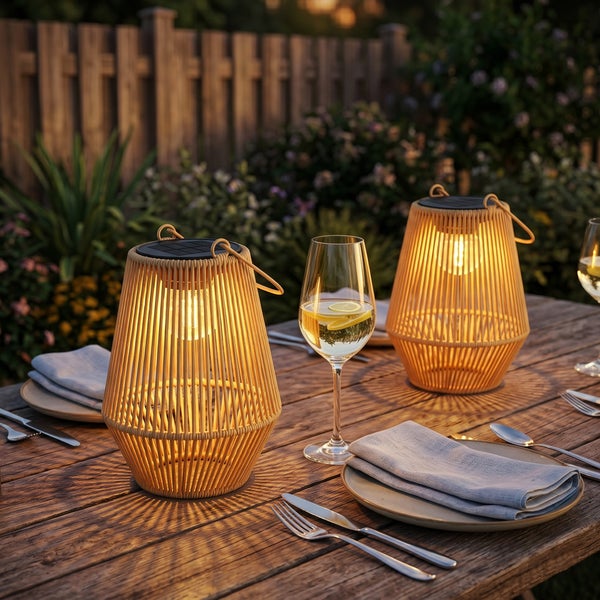 Zwei leuchtende Solar-Gartenlaternen im Korb-Design auf einem gedeckten Holztisch im Garten mit Pflanzen und einem Holzzaun.