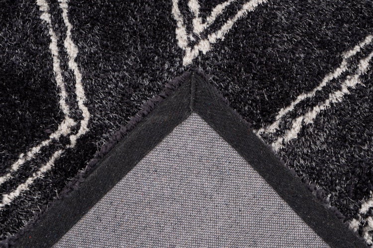 Detailansicht eines Hochflor-Teppichs mit weißem abstraktem Muster auf dunklem Grund sowie der gewebten Rückseite mit Textilkante.