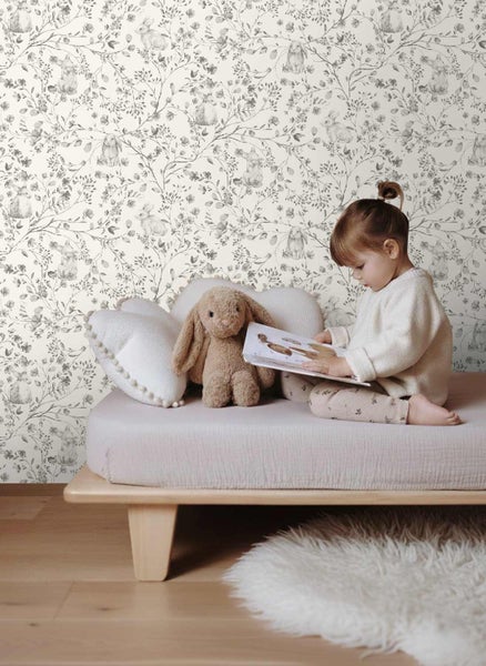 Kinderzimmer mit einer Vliestapete mit Hasen- und Blumenmuster, ein Kind sitzt auf einem niedrigen Holzbett und liest ein Buch.