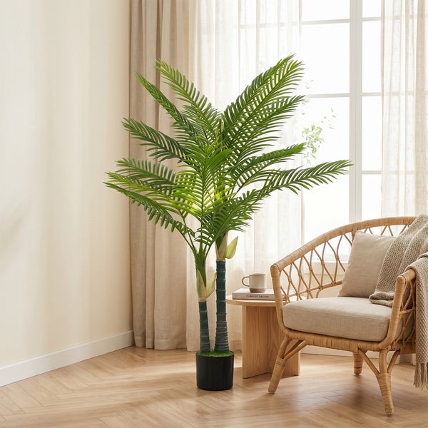 Künstliche Palme in schwarzem Topf in einem hellen Wohnzimmer neben einem Rattansessel und einem Beistelltisch aus Holz.
