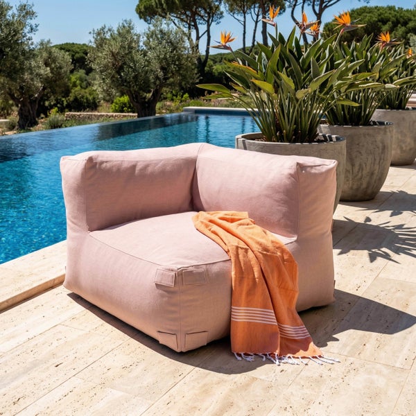 Moderner rosa Outdoor-Sitzsack mit orangefarbenem Handtuch auf einer Steinterrasse an einem Pool, umgeben von mediterranen Pflanzen.