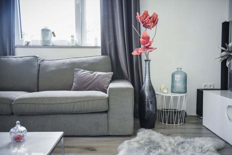 Modernes Wohnzimmer mit grauem Sofa, hoher Vase mit roten Kunstblumen, weißem Metalldraht-Beistelltisch und grauem Kunstfellteppich.