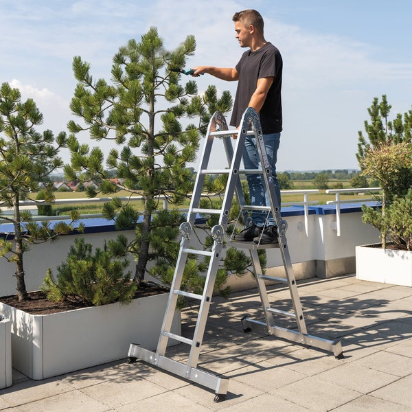 Ein Mann schneidet mit einer elektrischen Gartenschere einen Nadelbaum auf einer Dachterrasse, während er auf einer stabilen Aluminiumleiter steht.