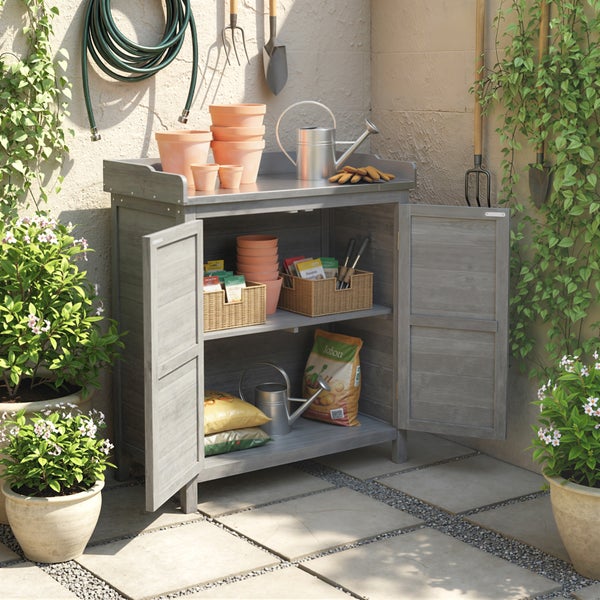 Grauer Garten-Pflanztisch aus Holz mit zwei Türen, Metall-Arbeitsfläche und Stauraum für Gartenwerkzeug und Töpfe im Außenbereich.