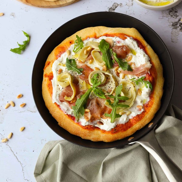 Pizza in einer Bratpfanne mit Mozzarella, Artischocken, Rucola, Schinken und Pinienkernen