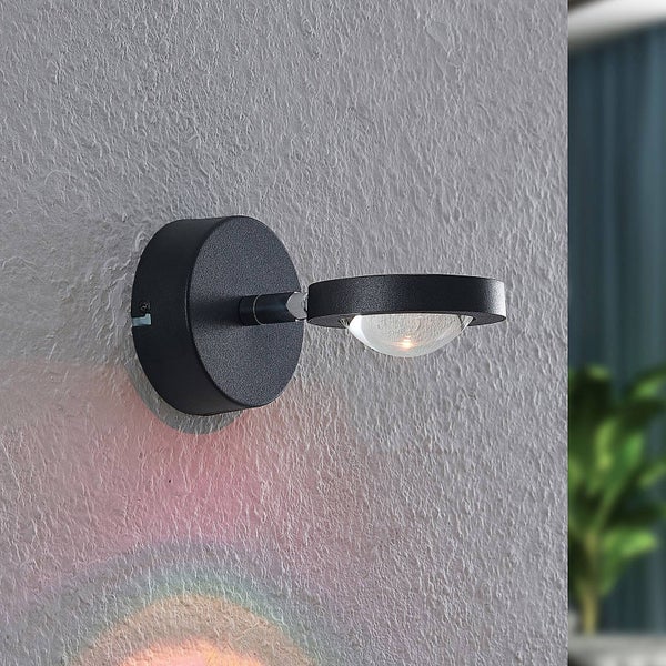 Moderne schwarze Wandleuchte mit rundem Sockel und ringförmigem Kopf, die einen Regenbogen-Lichteffekt auf eine strukturierte Wand wirft.
