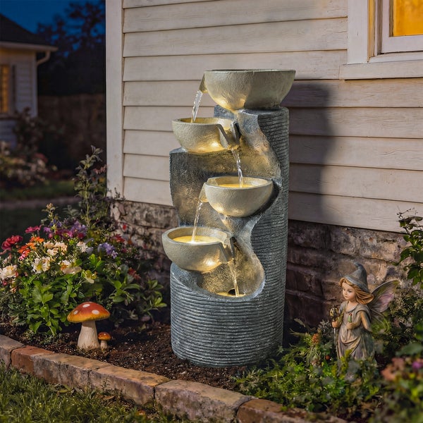 Beleuchteter Kaskadenbrunnen in Steinoptik mit vier Schalen in einem Gartenbeet vor einer Hauswand.