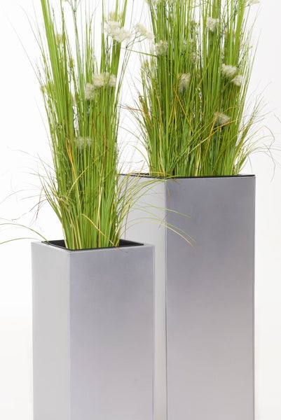 Zwei hohe silberfarbene rechteckige Pflanzkübel mit künstlichem grünen Gras und kleinen weißen Blüten.