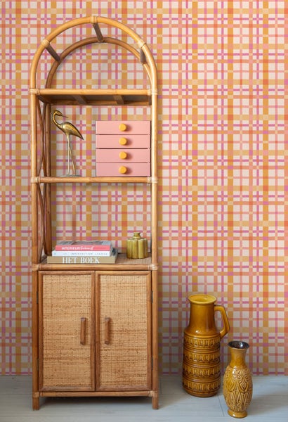 Karierte Tapete in Orange, Pink und Gelb hinter einem hohen Rattanregal mit Dekoration und Keramikvasen.
