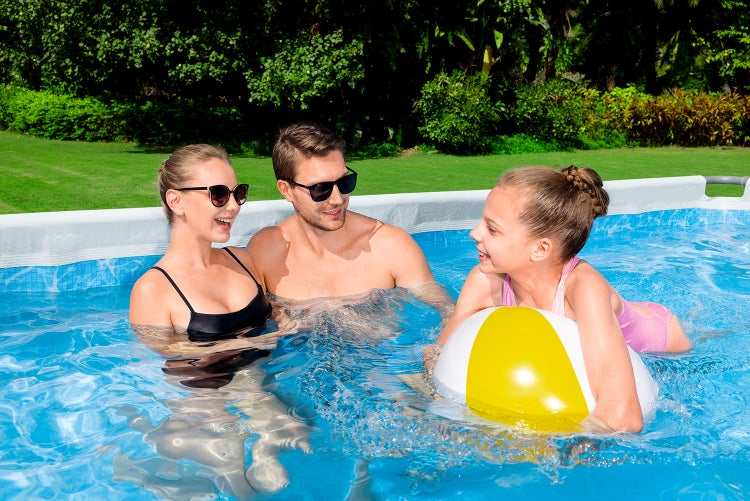 Familie mit Kind in einem rechteckigen Aufstellpool im Garten, das Kind spielt mit einem Wasserball.
