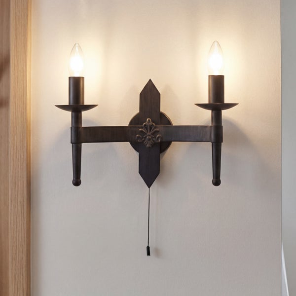 Zweiflammige Wandleuchte im rustikalen Design mit Zierornament und Zugschalter an einer Wand montiert.