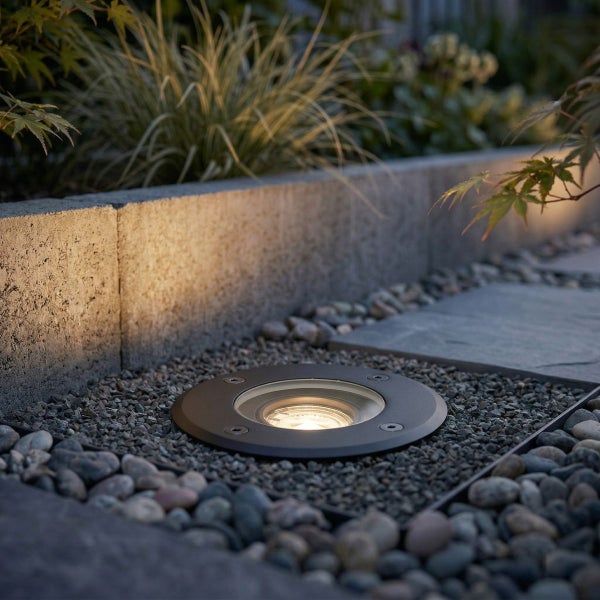 Bodeneinbaustrahler im Kiesbeet neben einer Gartenmauer aus Beton mit warmweißem Licht.