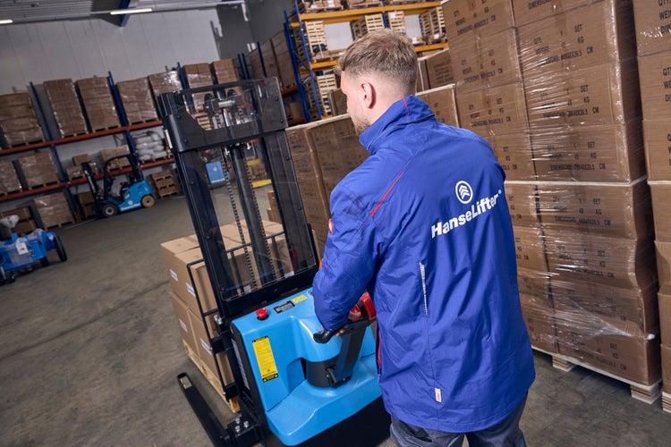 Ein Mitarbeiter bedient einen elektrischen HanseLifter Hochhubwagen in einem organisierten Lagerhaus mit vielen gestapelten Kartons.