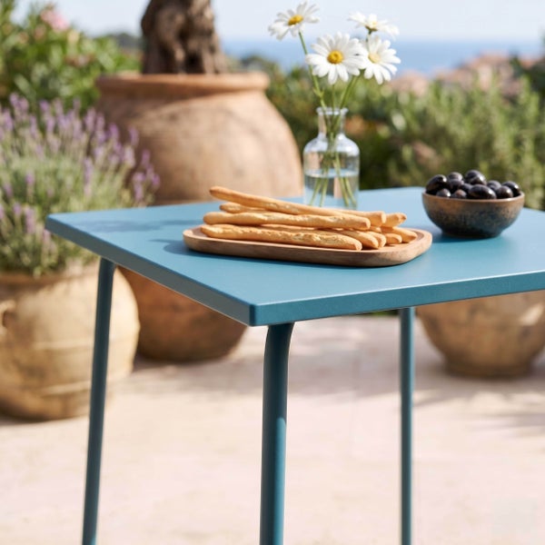 Quadratischer Gartentisch aus Metall auf einer sonnigen Terrasse mit Gebäckstangen, Oliven und Blumen in einer Vase.