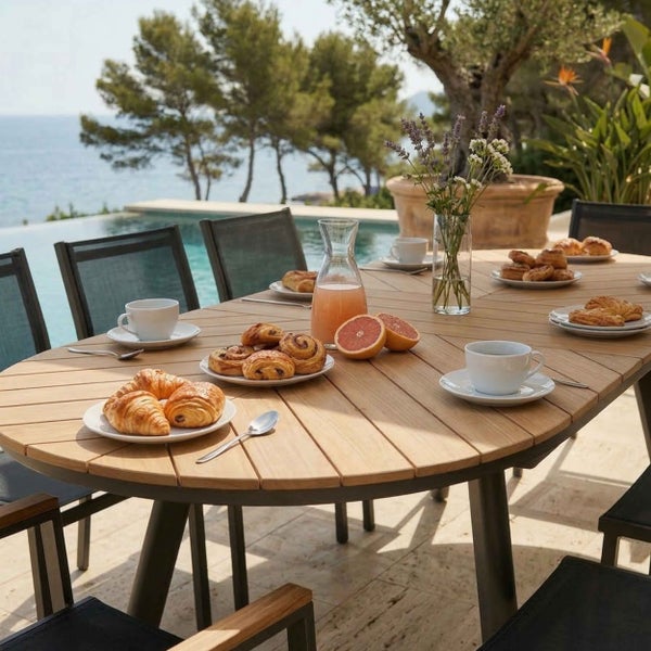 Gartentisch aus Holz mit Stühlen auf einer hellen Terrasse neben einem Pool, gedeckt mit Frühstück und Blumen vor einer Kulisse mit Meer und Bäumen.